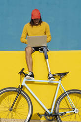 Young man with bicycle working on laptop while sitting on multi colored wall - JCZF00289