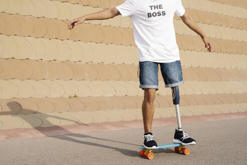 Junger Mann mit Bein- und Fußprothese steht auf einem Skateboard auf einem Sportplatz - JCZF00300