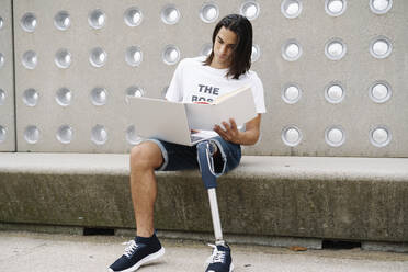 Man holding book while using laptop against wall - JCZF00307