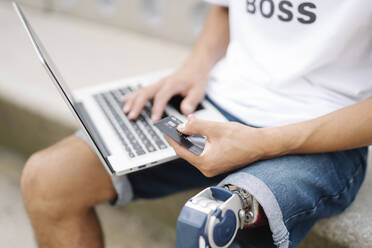 Man holding credit card while working on laptop outdoors - JCZF00309