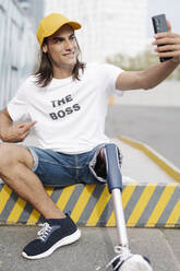 Mann mit Bein- und Fußprothese macht Selfie, während er auf der Straße in der Stadt sitzt - JCZF00319