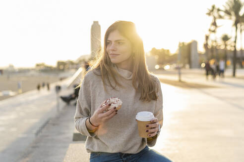 Nachdenkliche junge Frau, die einen frischen Donut mit einem Einwegbecher in der Hand hält, während sie an der Promenade sitzt und bei Sonnenuntergang wegschaut - AFVF07275