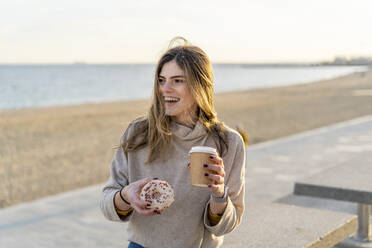 Fröhliche Frau, die einen frischen Donut mit einem Einwegbecher in der Hand hält, während sie am Strand sitzt und bei Sonnenuntergang in den Himmel schaut - AFVF07277