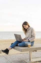 Schöne Frau, die einen Laptop benutzt, während sie mit einem Einwegbecher auf einer Bank an der Promenade am Meer sitzt, gegen den Himmel - AFVF07278