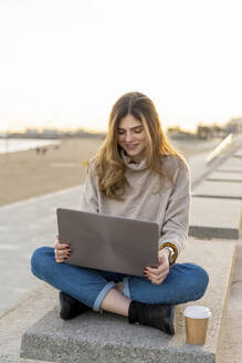 Lächelnde junge Frau sitzt im Schneidersitz mit Laptop und Einwegbecher auf einer Bank an der Promenade gegen den Himmel - AFVF07280