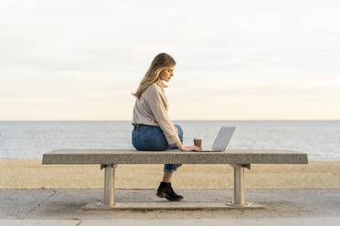 Junge Frau, die einen Laptop benutzt, während sie bei Sonnenuntergang auf einer Bank an der Promenade am Meer sitzt - AFVF07281