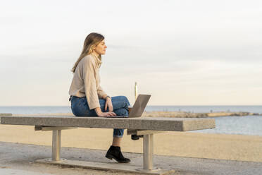 Nachdenkliche Frau, die mit ihrem Laptop auf einer Bank an der Strandpromenade gegen den Himmel bei Sonnenuntergang sitzt und wegschaut - AFVF07283