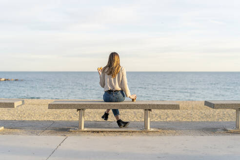 Junge Frau schaut auf das Meer, während sie auf einer Bank sitzt und den Sonnenuntergang genießt - AFVF07284
