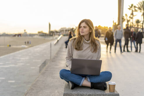 Nachdenkliche junge Frau, die im Schneidersitz mit Laptop und Einwegbecher auf einer Bank an der Promenade sitzt und wegschaut - AFVF07286