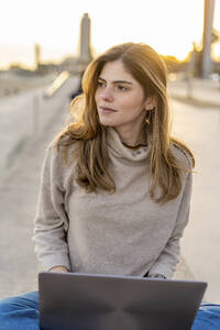Schöne junge Frau, die wegschaut, während sie mit einem Laptop an der Promenade bei Sonnenuntergang sitzt - AFVF07287