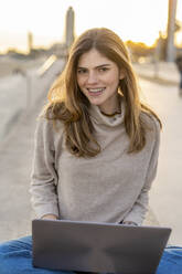 Lächelnde schöne junge Frau sitzt mit Laptop an der Strandpromenade bei Sonnenuntergang - AFVF07288