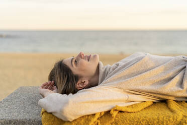 Junge Frau, die auf einer Bank an der Strandpromenade liegt und nach oben schaut, während die Sonne untergeht - AFVF07292