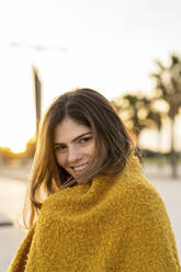 Lächelnde schöne junge Frau in brauner Jacke am Strand bei Sonnenuntergang eingewickelt - AFVF07293