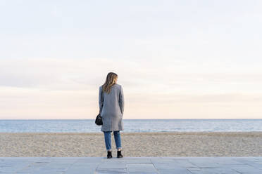 Junge Frau schaut auf das Meer, während sie auf der Promenade am Strand steht und den Sonnenuntergang genießt - AFVF07295