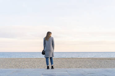 Young woman looking at sea while standing on promenade at beach enjoying sunset - AFVF07295