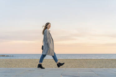 Happy beautiful woman walking on promenade at beach while looking away against sky during sunset - AFVF07297