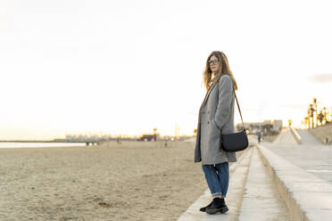 Nachdenkliche junge Frau mit Hand in der Tasche, die auf der Strandpromenade steht und bei Sonnenuntergang wegschaut - AFVF07299