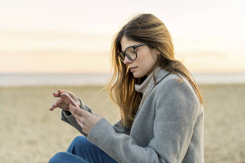 Schöne junge Frau mit langen braunen Haaren verwendet Smartphone am Strand bei Sonnenuntergang - AFVF07300