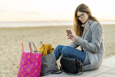 Schöne junge Frau, die ihr Smartphone benutzt, während sie mit Einkaufstaschen am Strand bei Sonnenuntergang sitzt - AFVF07301