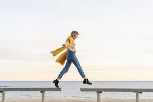 Cheerful young woman jumping from bench to another at beach promenade against sky during sunset - AFVF07305