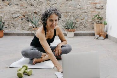 Smiling woman using laptop for learning yoga through online tutorial while sitting at back yard - TCEF01188