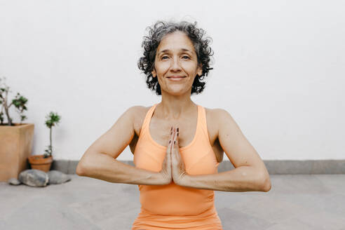 Lächelnde Frau übt Yoga mit verschränkten Händen an einer weißen Wand im Hinterhof - TCEF01191
