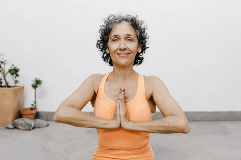 Smiling woman practicing yoga with hands clasped against white wall at back yard - TCEF01191