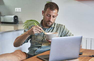Entspannter Freiberufler isst Nudeln, während er mit seinem Laptop am Esstisch sitzt - VEGF02978
