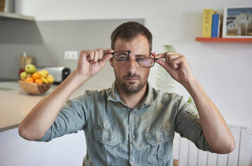 Mittlerer Erwachsener, der eine zerbrochene Brille hält, während er zu Hause sitzt - VEGF02983