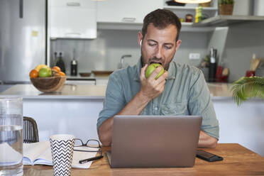 Handsome male freelancer eating fresh apple while working at home with laptop - VEGF02986