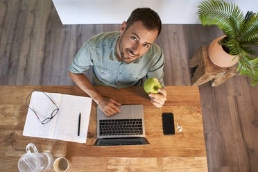 Smiling businessman holding fresh apple while working at home - VEGF02989