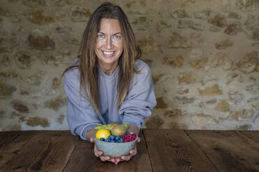 Smiling female nutritionist with various fruits in bowl sitting at table against stone wall - DLTSF01265