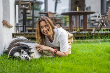 Glückliche Frau kniet neben einem Hund und entspannt sich im Gras im Hinterhof - DLTSF01266