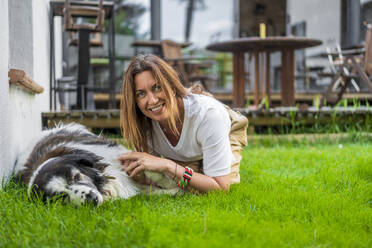 Happy woman kneeling by dog relaxing on grass at back yard - DLTSF01266