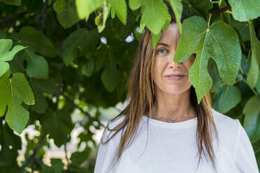 Mature woman with blue eyes standing under green leaves at back yard - DLTSF01268