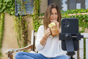 Ernährungsberaterin, die Saft aus einem Glas trinkt, während sie mit ihrem Smartphone im Hinterhof vloggt - DLTSF01273