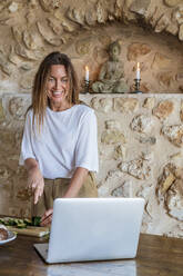 Smiling female nutritionist vlogging while cutting cucumber against stone wall - DLTSF01277