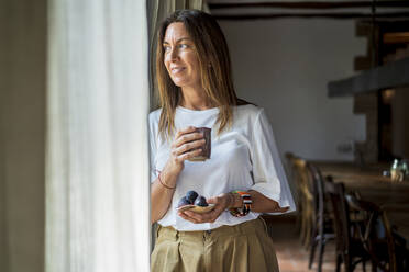 Thoughtful woman holding drink and fruits while looking through window - DLTSF01285