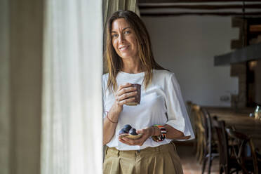 Smiling woman holding drink and fruits while looking through window - DLTSF01286