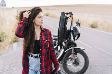 Female biker with hand in hair looking away while standing on road - DAMF00511