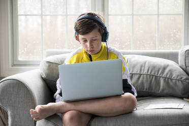 Junge mit Laptop für virtuelle Schule sitzt auf der Couch im Wohnzimmer - CAVF89735