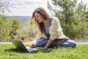 Smiling woman using laptop while sitting on grass at backyard - DLTSF01297