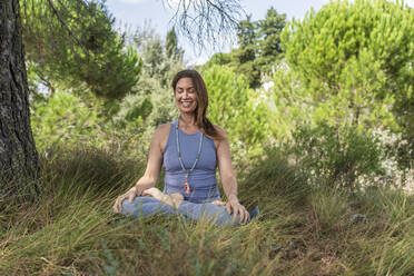 Lächelnde Frau übt Yoga, während sie unter einem Baum im Gras sitzt - DLTSF01299