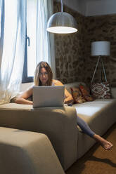 Woman working on laptop while sitting on sofa at home - DLTSF01301