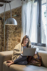 Woman working on laptop while sitting on sofa by window at home - DLTSF01303