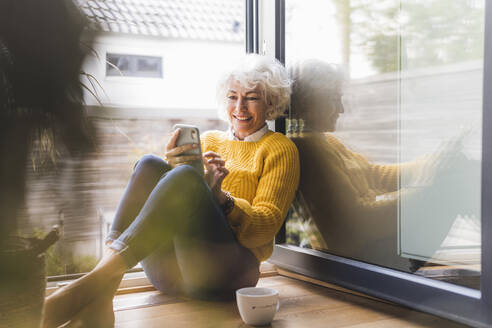 Lächelnde Frau, die zu Hause sitzend eine Textnachricht auf ihrem Smartphone schreibt - UUF21613