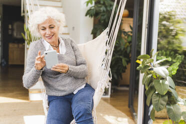Ältere Frau mit Smartphone auf einer Schaukel sitzend zu Hause - UUF21633