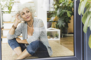 Mature woman looking through window with head in hands while sitting at home - UUF21636