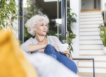 Nachdenkliche reife Frau, die Kaffee trinkt und wegschaut, während sie zu Hause auf der Couch sitzt - UUF21651