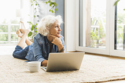 Mature woman lying on front while looking away working on laptop at home stock photo